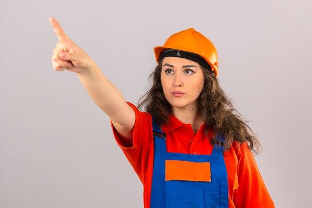 Mujer joven constructor en uniforme de construcción y casco de seguridad apuntando a algún lugar con el dedo mirando seriamente sobre la pared blanca aislada