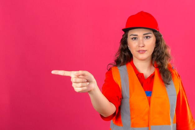 Mujer joven constructor en uniforme de construcción y casco de seguridad apuntando con el dedo hacia el lado sobre la pared rosa aislada