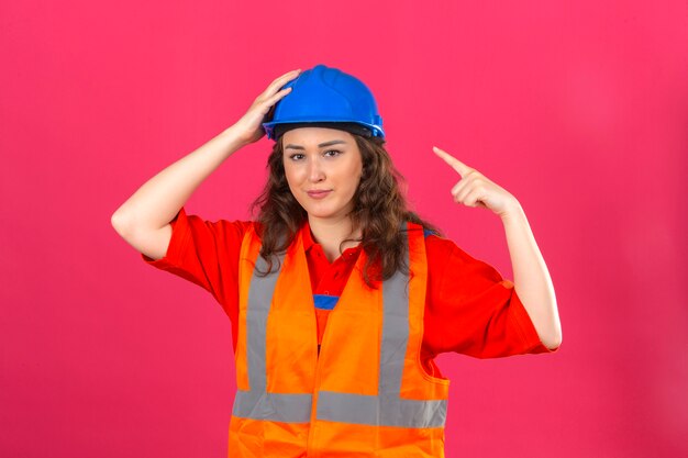 Mujer joven constructor en uniforme de construcción y casco de seguridad y apuntando a su casco con el dedo índice sobre la pared rosa aislada