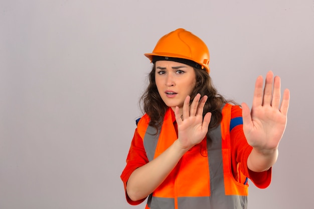 Mujer joven constructor en uniforme de construcción y casco de seguridad asustado y aterrorizado con expresión de miedo detener el gesto con las manos sobre la pared blanca aislada