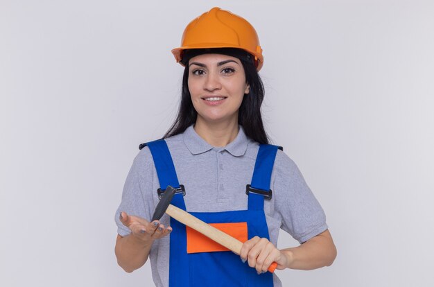 Mujer joven constructor en uniforme de construcción y casco de seguridad balanceando un martillo mirando al frente sonriendo confiado de pie sobre la pared blanca