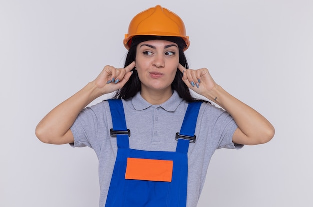 Mujer joven constructor en uniforme de construcción y casco de seguridad cubriendo las orejas con los dedos mirando confundido parado sobre pared blanca