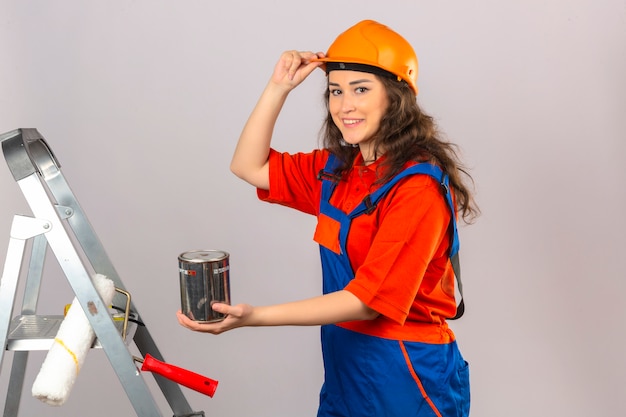 Mujer joven constructor en uniforme de construcción y casco de seguridad en una escalera de metal con pintura puede sonreír y tocar su casco sobre pared blanca aislada