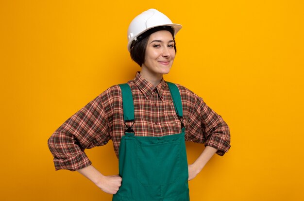 Mujer joven constructor en uniforme de construcción y casco de seguridad feliz y positivo sonriendo alegremente con las manos en la cadera de pie en naranja