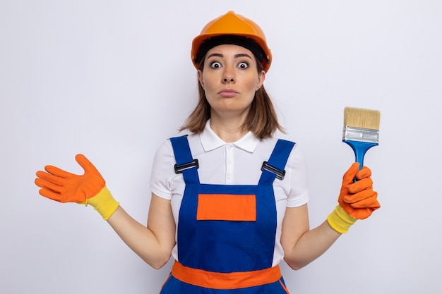 Mujer joven constructor en uniforme de construcción y casco de seguridad en guantes de goma con pincel confundido encogiéndose de hombros de pie sobre la pared blanca