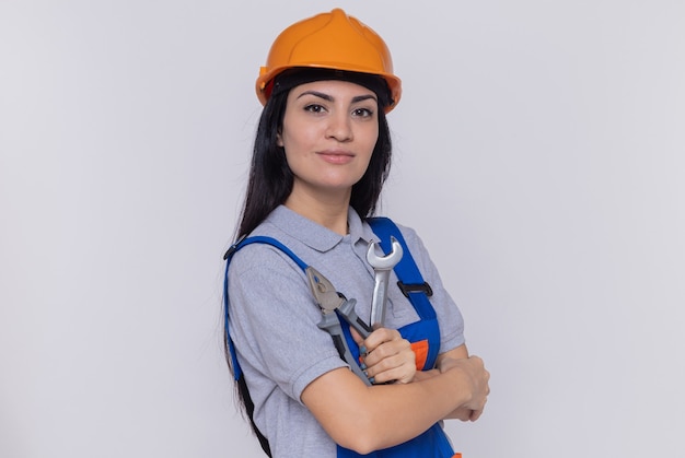 Mujer joven constructor en uniforme de construcción y casco de seguridad con llave y alicates mirando al frente sonriendo confiado de pie sobre la pared blanca