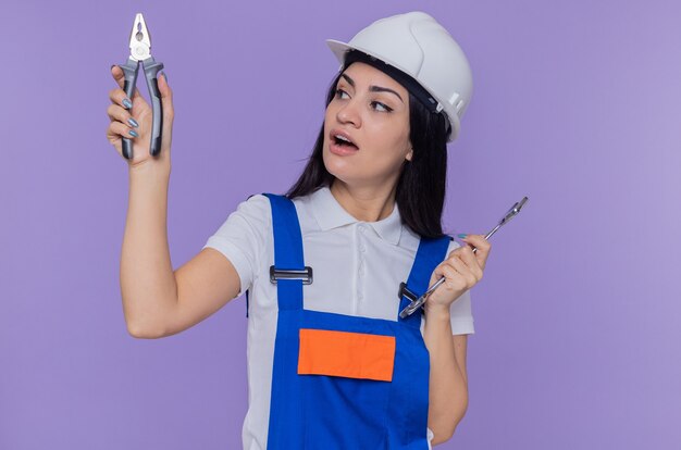 Mujer joven constructor en uniforme de construcción y casco de seguridad con llave y alicates que parece confundido tratando de tomar una decisión de pie sobre la pared púrpura