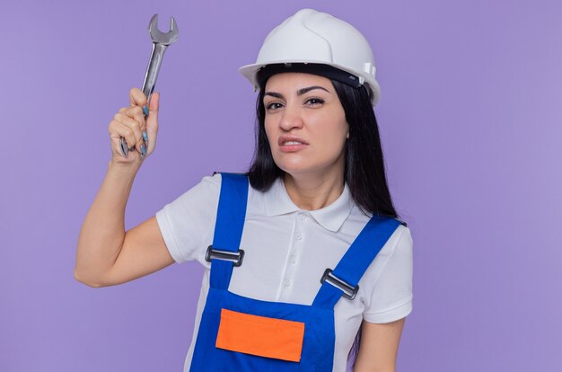 Mujer joven constructor en uniforme de construcción y casco de seguridad con llave mirando al frente con expresión escéptica de pie sobre la pared púrpura