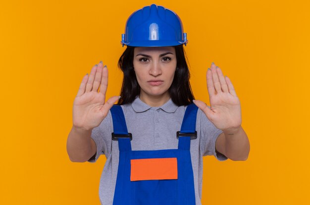 Mujer joven constructor en uniforme de construcción y casco de seguridad mirando al frente con cara seria haciendo gesto de parada con las manos de pie sobre la pared naranja