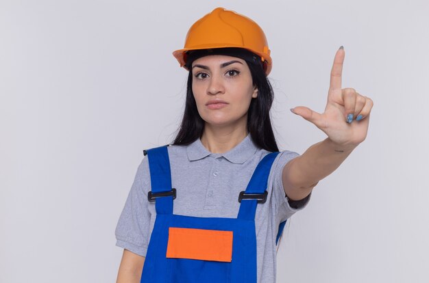 Mujer joven constructor en uniforme de construcción y casco de seguridad mirando al frente con cara seria que muestra la advertencia del dedo índice de pie sobre la pared blanca