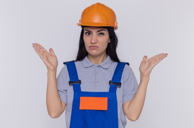 Mujer joven constructor en uniforme de construcción y casco de seguridad mirando al frente confundido y muy ansioso con los brazos levantados de pie sobre la pared blanca