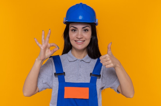 Mujer joven constructor en uniforme de construcción y casco de seguridad mirando al frente feliz y positivo mostrando el pulgar hacia arriba y el signo de ok de pie sobre la pared naranja