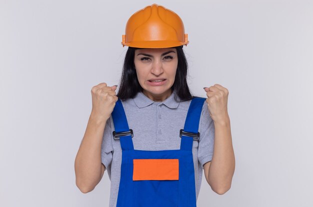 Mujer joven constructor en uniforme de construcción y casco de seguridad mirando al frente con los puños cerrados, enojado y frustrado de pie sobre la pared blanca