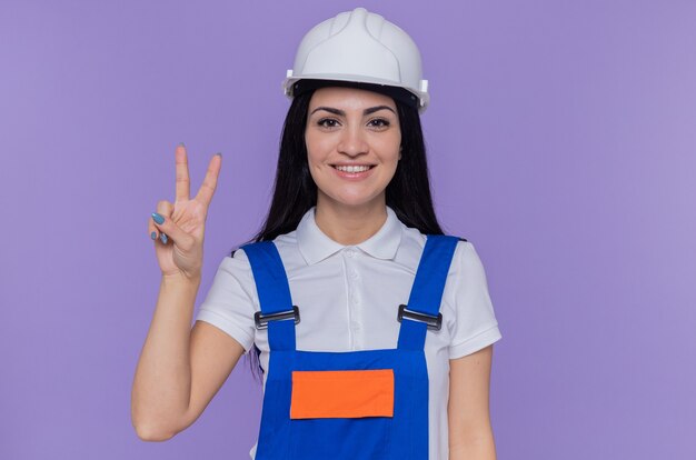 Mujer joven constructor en uniforme de construcción y casco de seguridad mirando al frente sonriendo alegremente mostrando v-sign de pie sobre la pared púrpura