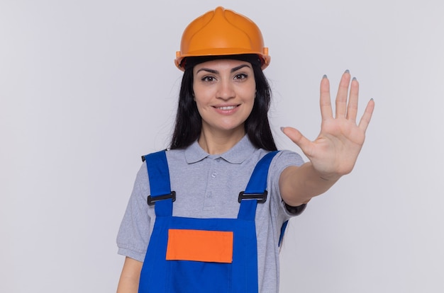 Mujer joven constructor en uniforme de construcción y casco de seguridad mirando al frente sonriendo confiado haciendo gesto de parada con la mano abierta de pie sobre la pared blanca