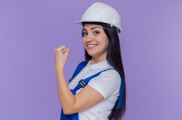Mujer joven constructor en uniforme de construcción y casco de seguridad mirando al frente sonriendo confiado mostrando puño parado sobre la pared púrpura