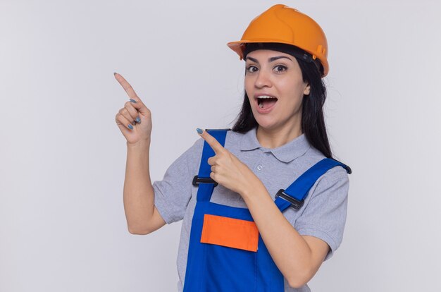 Mujer joven constructor en uniforme de construcción y casco de seguridad mirando al frente sonriendo feliz y positivo apuntando con los dedos índices al lado de pie sobre la pared blanca