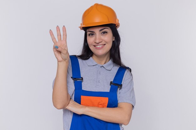 Mujer joven constructor en uniforme de construcción y casco de seguridad mirando al frente sonriendo mostrando el número tres con los dedos de pie sobre la pared blanca