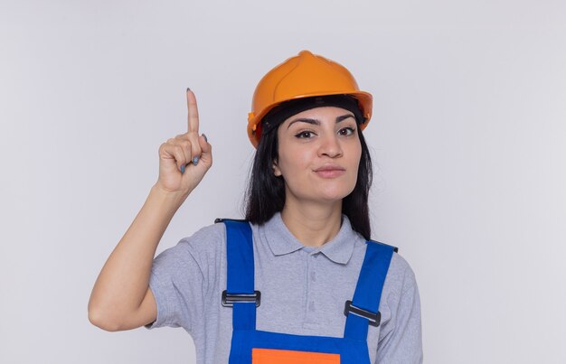 Mujer joven constructor en uniforme de construcción y casco de seguridad mirando al frente con una sonrisa en la cara inteligente que muestra el dedo índice de pie sobre la pared blanca