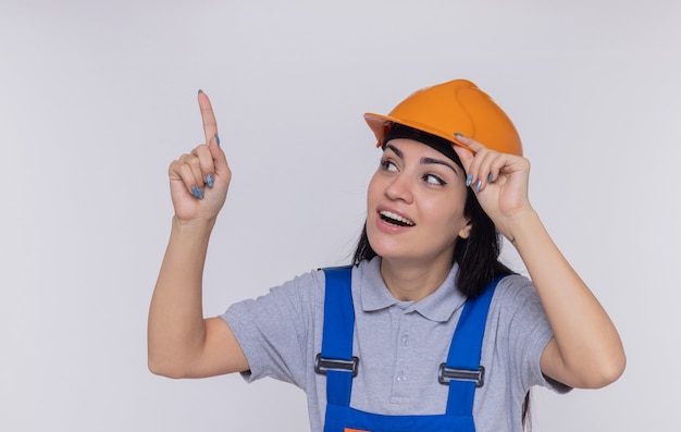 Mujer joven constructor en uniforme de construcción y casco de seguridad mirando hacia arriba sonriendo apuntando con el dedo índice a algo