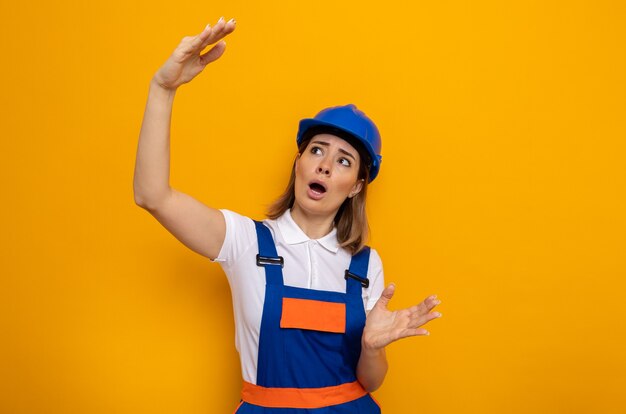 Mujer joven constructor en uniforme de construcción y casco de seguridad mirando asombrado y sorprendido haciendo gesto de tamaño con las manos de pie sobre la pared naranja