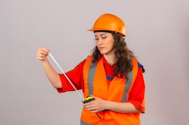 Mujer joven constructor en uniforme de construcción y casco de seguridad mirando cinta métrica con cara seria sobre pared blanca aislada
