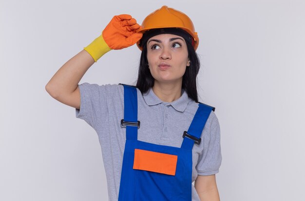 Mujer joven constructor en uniforme de construcción y casco de seguridad mirando desconcertado tocando su casco de pie sobre la pared blanca