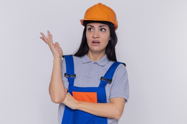 Mujer joven constructor en uniforme de construcción y casco de seguridad mirando a un lado confundido y disgustado con el brazo extendido de pie sobre la pared blanca