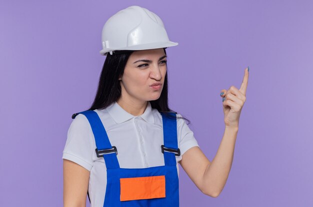 Mujer joven constructor en uniforme de construcción y casco de seguridad mirando a un lado confundido y disgustado con el brazo levantado