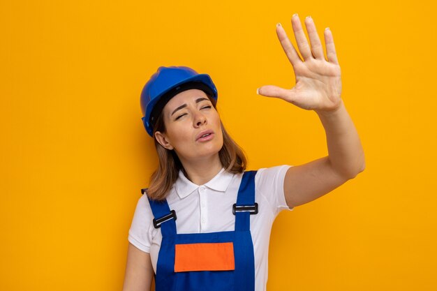 Mujer joven constructor en uniforme de construcción y casco de seguridad mirando a un lado entrecerrar los ojos y levantar el brazo de pie en naranja