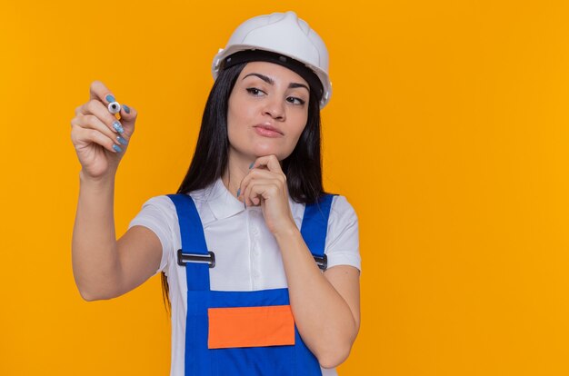 Mujer joven constructor en uniforme de construcción y casco de seguridad mirando a un lado con expresión pensativa escribiendo algo con bolígrafo de pie sobre la pared naranja