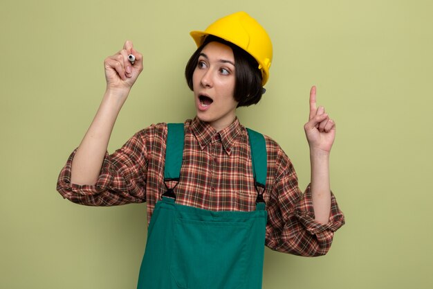 Mujer joven constructor en uniforme de construcción y casco de seguridad mirando a un lado feliz y sorprendido escribiendo en el aire con la pluma de pie en verde