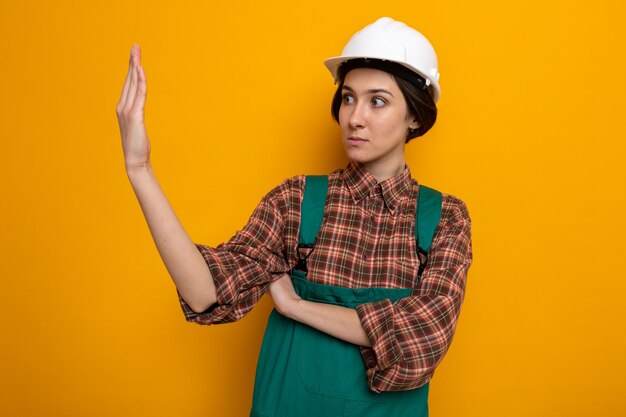 Mujer joven constructor en uniforme de construcción y casco de seguridad mirando a un lado a su mano preocupada y confundida de pie en naranja