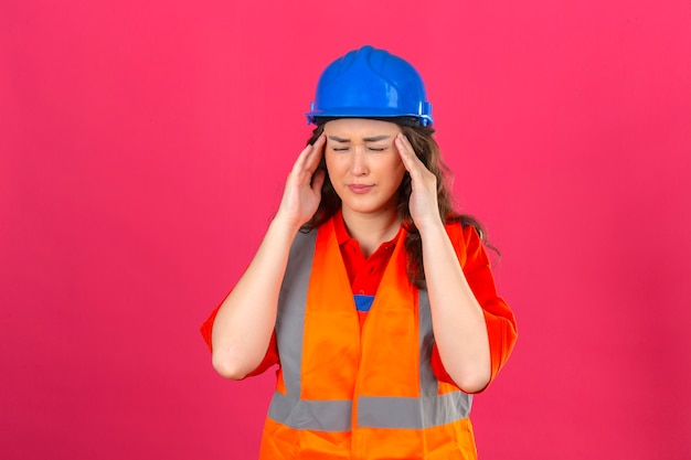 Mujer joven constructor en uniforme de construcción y casco de seguridad mirando mal tocando su cabeza con fuerte dolor de cabeza sobre pared rosa aislada