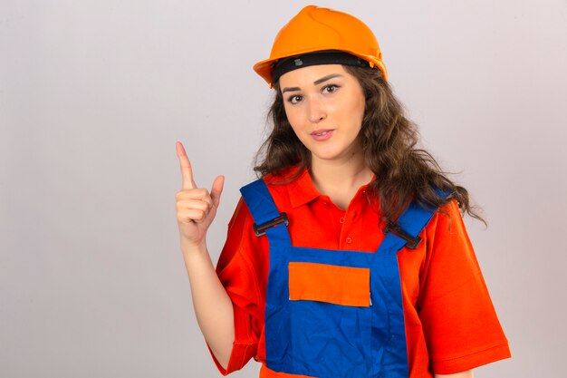 Mujer joven constructor en uniforme de construcción y casco de seguridad mirando seguro dedo apuntando hacia arriba sobre pared blanca