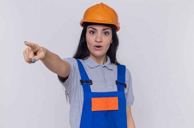 Mujer joven constructor en uniforme de construcción y casco de seguridad mirando sorprendido apuntando con el dedo índice a algo parado sobre la pared blanca