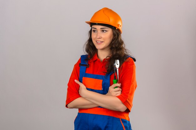 Mujer joven constructor en uniforme de construcción y casco de seguridad de pie con los brazos cruzados con herramientas de construcción mirando a otro lado con una sonrisa sobre la pared blanca aislada