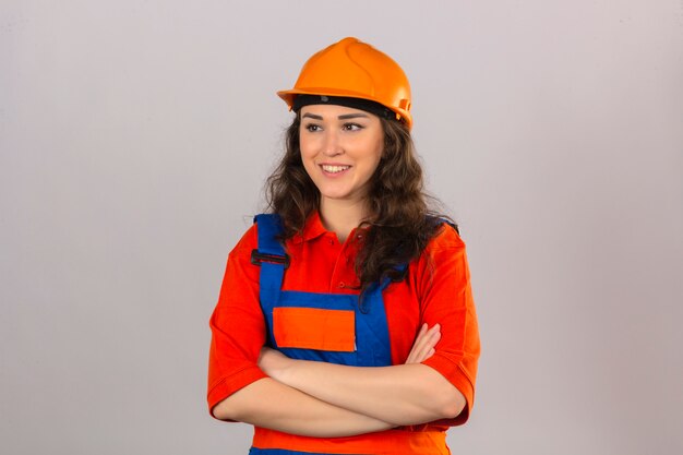 Mujer joven constructor en uniforme de construcción y casco de seguridad de pie con los brazos cruzados sonriendo amable sobre pared blanca aislada