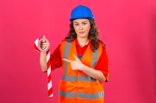 Mujer joven constructor en uniforme de construcción y casco de seguridad de pie con cinta adhesiva y apuntando con el dedo índice frunciendo el ceño sobre la pared rosada aislada
