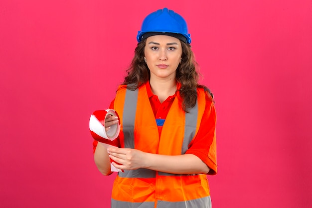Mujer joven constructor en uniforme de construcción y casco de seguridad de pie con cinta adhesiva con cara seria sobre wall_ rosa aislado