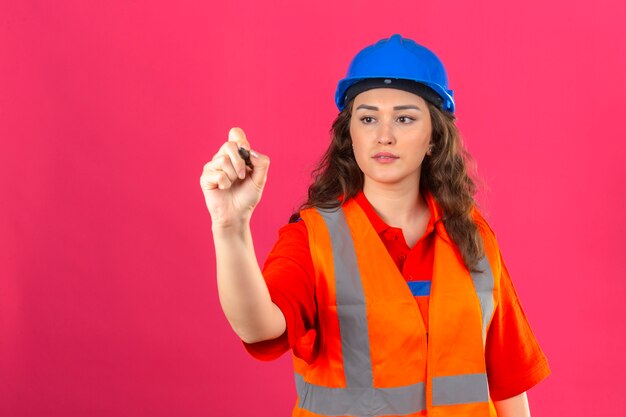 Mujer joven constructor en uniforme de construcción y casco de seguridad de pie escribiendo en el aire con lápiz sobre pared rosa aislado