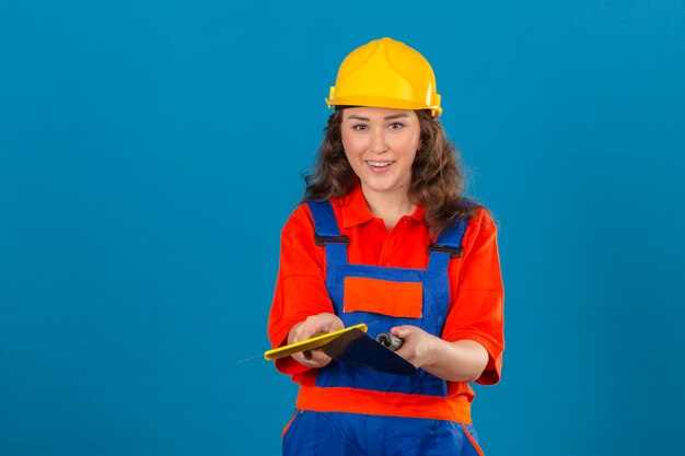 Mujer joven constructor en uniforme de construcción y casco de seguridad de pie con espátula sonriendo amigable sobre pared azul aislado