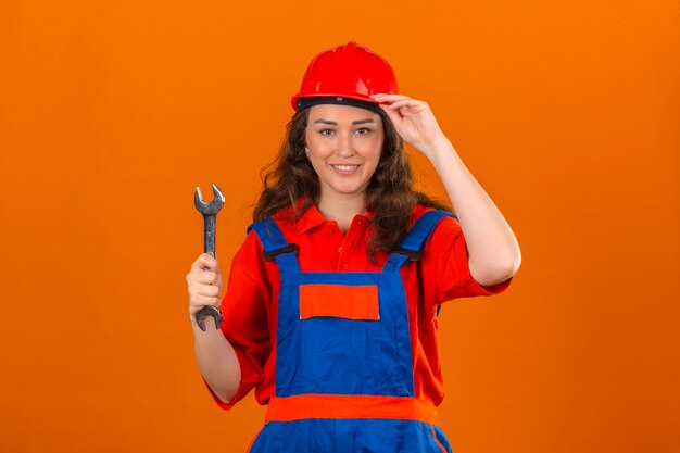 Mujer joven constructor en uniforme de construcción y casco de seguridad de pie con una llave tocando su casco sonriendo sobre pared naranja aislada