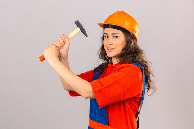 Mujer joven constructor en uniforme de construcción y casco de seguridad de pie con un martillo que amenaza con golpearlo sobre una pared blanca aislada