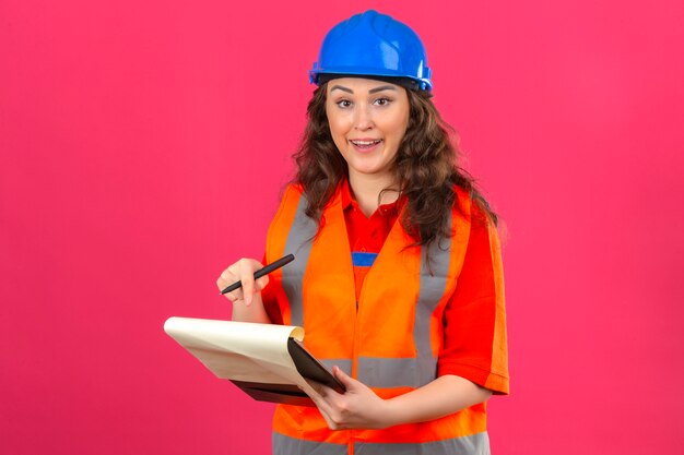 Mujer joven constructor en uniforme de construcción y casco de seguridad de pie con portapapeles apuntando sobre él disgustado sobre pared rosa aislado