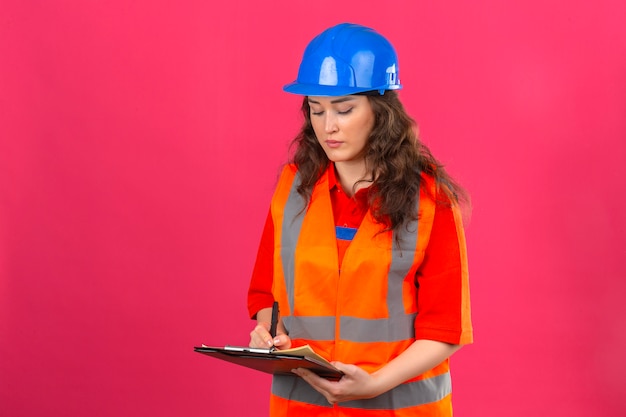 Mujer joven constructor en uniforme de construcción y casco de seguridad de pie con portapapeles haciendo notas con cara seria sobre pared rosa aislado