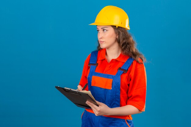 Mujer joven constructor en uniforme de construcción y casco de seguridad de pie con portapapeles haciendo notas en él sobre la pared azul aislada