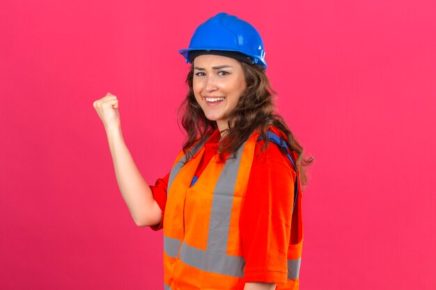 Mujer joven constructor en uniforme de construcción y casco de seguridad de pie con el puño levantado sonriendo con cara feliz concepto ganador sobre pared rosa aislado