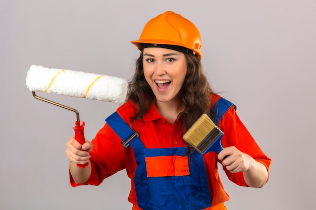 Mujer joven constructor en uniforme de construcción y casco de seguridad de pie con rodillo de pintura y pincel sonriendo alegremente sobre pared blanca aislada