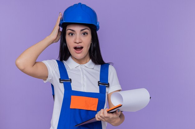 Mujer joven constructor en uniforme de construcción y casco de seguridad con portapapeles y lápiz mirando al frente asombrado y sorprendido de pie sobre la pared púrpura